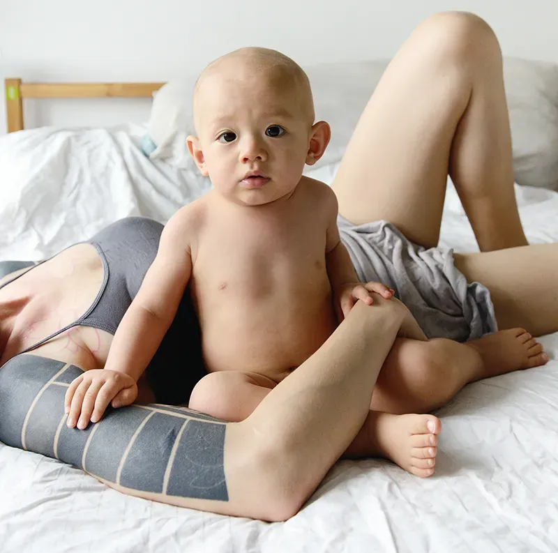 A young child seated with its mother lying down and holding them on the bed.