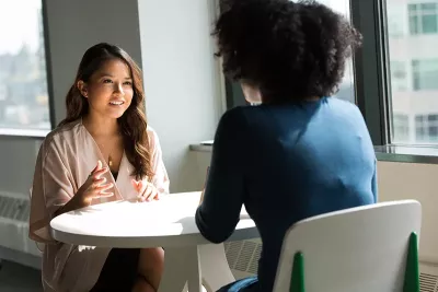two women sit at a round table talking to each other.