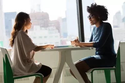 two women sit at a round table, talking to each other.