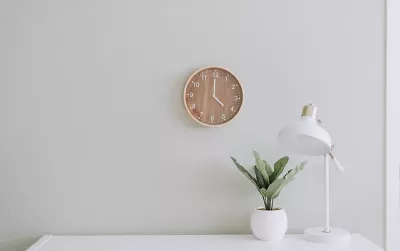 a wooden clock on a white wall next to a desk and plant.