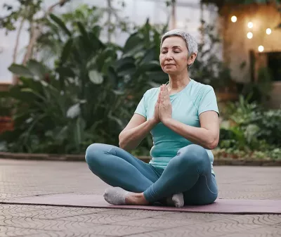 older woman with short hair seated criss-cross on a mat with her hands at heart center.