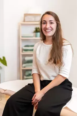 A female massage therapist sits on her massage table smiling for a photo.