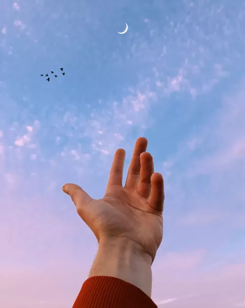 A hand reaches toward a sliver of moon in the sky.