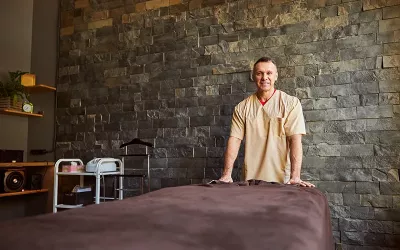 A male massage therapist stands at the head of a massage table, smiling into the camera.