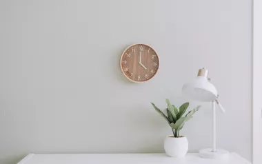 a wooden clock on a white wall next to a desk and plant.