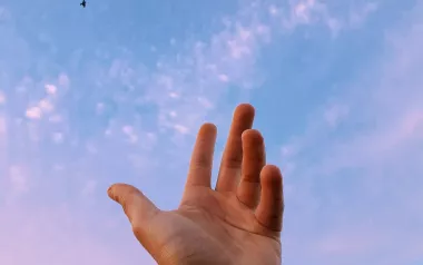 A hand reaches toward a sliver of moon in the sky.