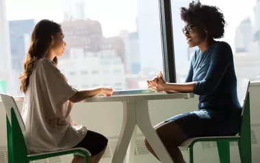 two women seated at a table across from each other, talking.