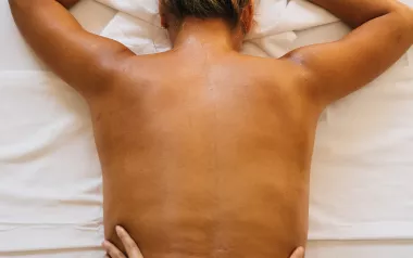 A woman lies face down on a massage table with her upper back exposed as an MT's hands work on her lower back.