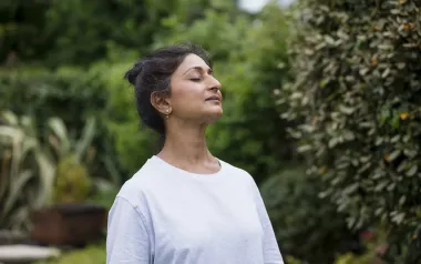 A woman with brown hair in a plain sleeved shirt takes a big deep breath with her eyes closed.