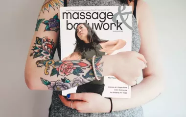 A woman with tattoos on her arms holding a copy of Massage & Bodywork July/August 2023.