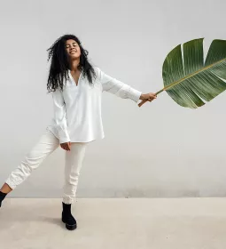 A woman balances on one leg while holding a giant leaf in the other hand, almost as a counterbalance to her raised leg.