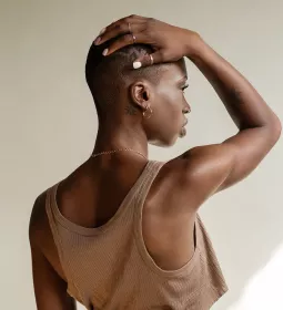 a Black woman in a tank top with a buzz cut holds her arm over her head, rotating her scapula.
