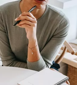 A woman in a long-sleeve shirt holds a black pen to her mouth as she thinks.