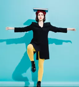 a woman in black and yellow clothes balances a book on her head while standing on one foot.