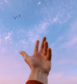 A hand reaches toward a sliver of moon in the sky.