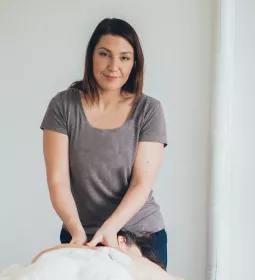 A female MT works on a prone client on a table. The MT is looking at the camera.