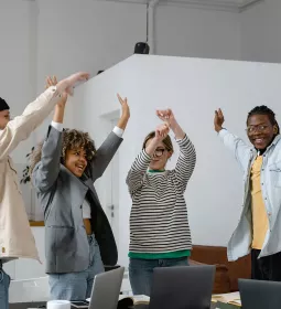 4 people of different ethnicities in a classroom celebrate with their arms in the air.