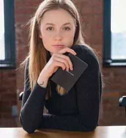 A woman holds a notepad and stares directly into the camera while sitting at a wood table.