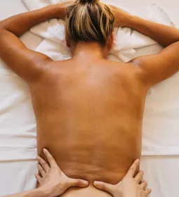 A woman lies face down on a massage table with her upper back exposed as an MT's hands work on her lower back.