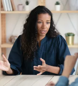 a woman with curly hair in a sweater sitting at a desk gestures with her hands.