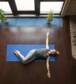 A woman lies on a blue yoga mat on the floor with her arms outstretched.