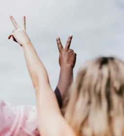 One person with white skin tone and another with dark skin tone make peace signs with their hands.