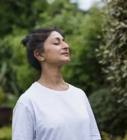 A woman with brown hair in a plain sleeved shirt takes a big deep breath with her eyes closed.