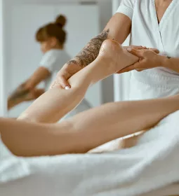 A massage therapist dressed in white works on a prone client's lower calf.