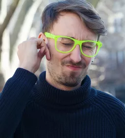 A man in a navy sewater and neon glasses clutches his ear and winces.