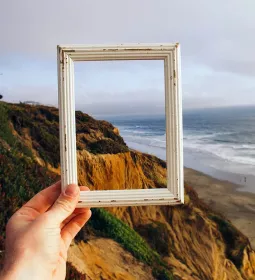 Hand holding small picture frame with ocean and cliffs in the background.