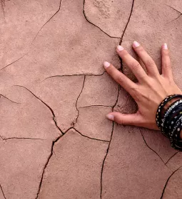 A hand with multiple bracelets touches cracked, dried dirt.