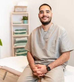 A massage therapist sits on the edge of a massage table, smiling for a photo.