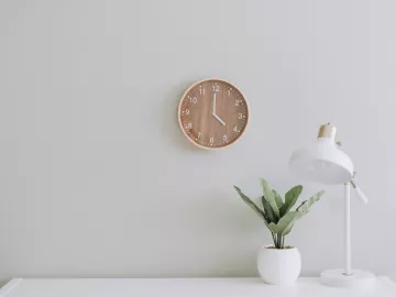 a wooden clock on a white wall next to a desk and plant.