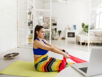 a woman on a yoga mat uses a stretching band on her feet as she follows an online class.