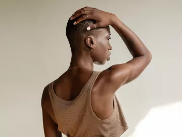 a Black woman in a tank top with a buzz cut holds her arm over her head, rotating her scapula.