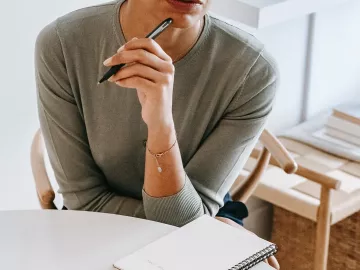 A woman in a long-sleeve shirt holds a black pen to her mouth as she thinks.