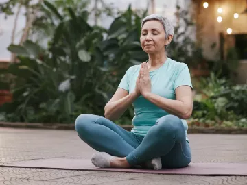 older woman with short hair seated criss-cross on a mat with her hands at heart center.