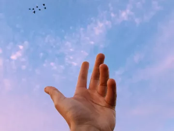 A hand reaches toward a sliver of moon in the sky.