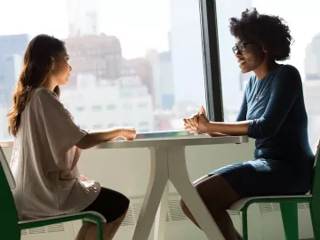 two women seated at a table across from each other, talking.