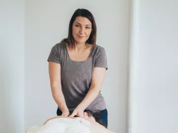A female MT works on a prone client on a table. The MT is looking at the camera. 