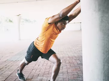 A Black man in exercise clothes pushes against a concrete column to stretch.