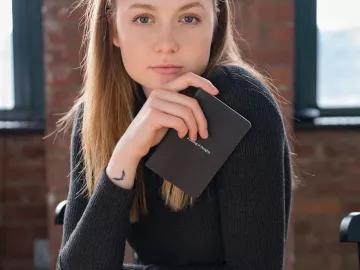 A woman holds a notepad and stares directly into the camera while sitting at a wood table.