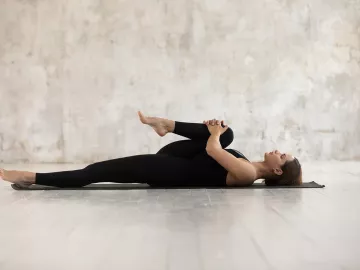 A woman on her back on a yoga mat stretches her right leg and glutes.
