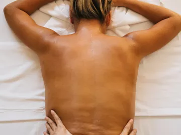 A woman lies face down on a massage table with her upper back exposed as an MT's hands work on her lower back.