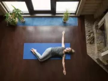 A woman lies on a blue yoga mat on the floor with her arms outstretched.