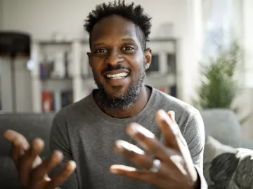 A Black man smiles and gestures largely with both hands.