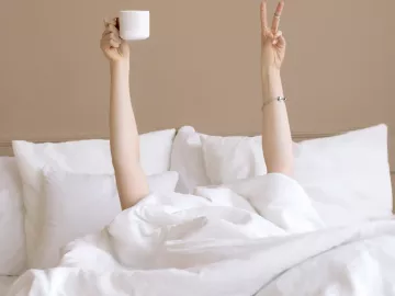 A person under the covers in bed holds up a peace sign with one hand and a coffee cup in the other hand.