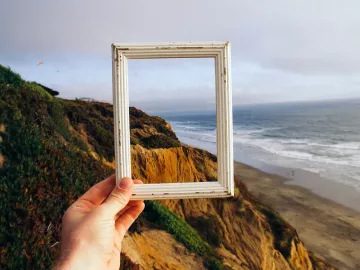 Hand holding small picture frame with ocean and cliffs in the background.