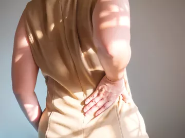 A person in a beige dress holds a hand to their lower back.