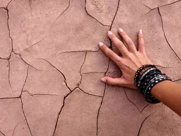 A hand with multiple bracelets touches cracked, dried dirt.